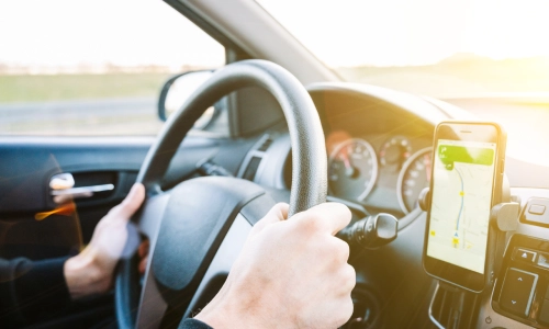 A driver holding a steering wheel with a GPS navigation app open on a smartphone mounted on the dashboard, representing rideshare services like Uber and potential accidents involving them.