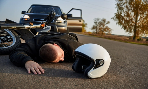 A motorcyclist lies on the asphalt beside a helmet, near a motorcycle and a car.