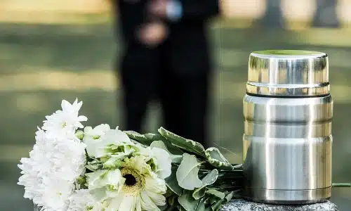 A wrongful death victim's urn on top of a grave next to flowers while a couple mourns in the background.