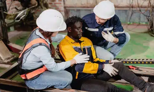 Two hardhat workers assisting a third injured coworker with their knee after a workplace accident.