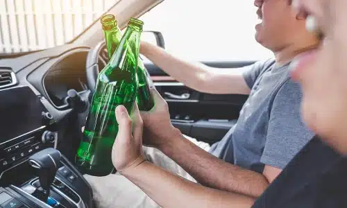 Two men who appear intoxicated hold bottles of alcohol while sitting inside a vehicle.